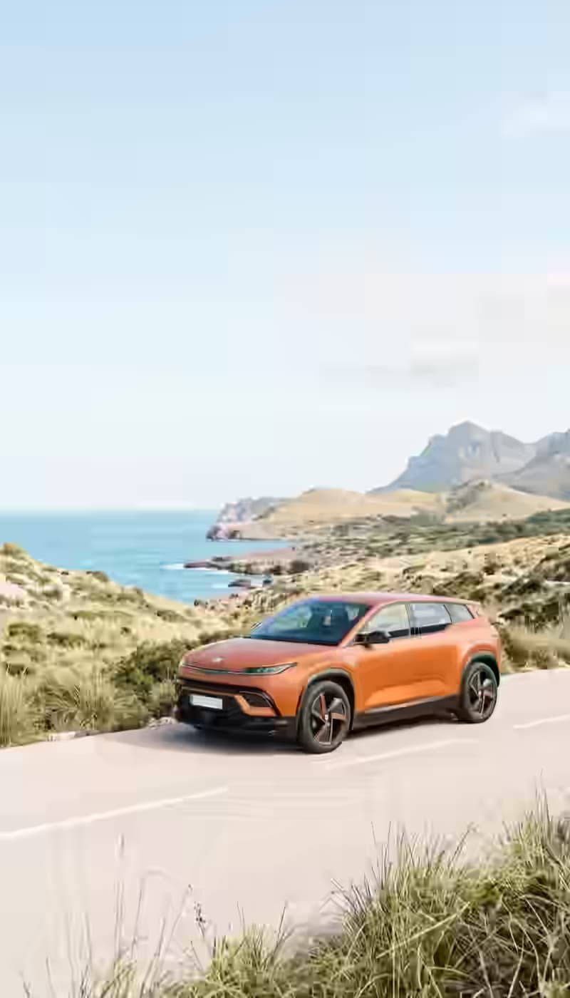 A scenic coastal road featuring a modern orange electric SUV from karpadu rent a car, driving through the mountainous landscape of Heraklion, Crete, with the Aegean Sea in the background under a clear sky.