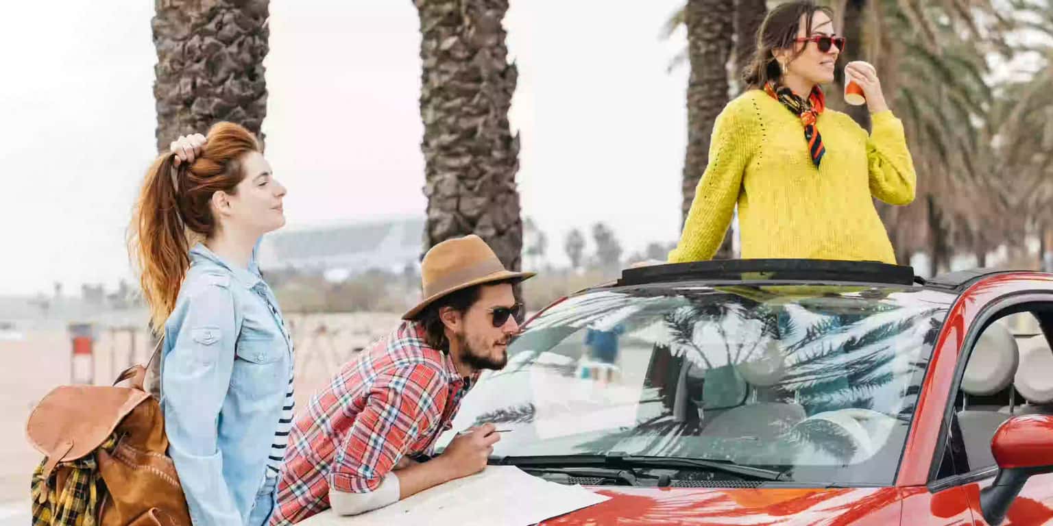 Three friends planning a road trip, looking at a map on the hood of a red convertible car.